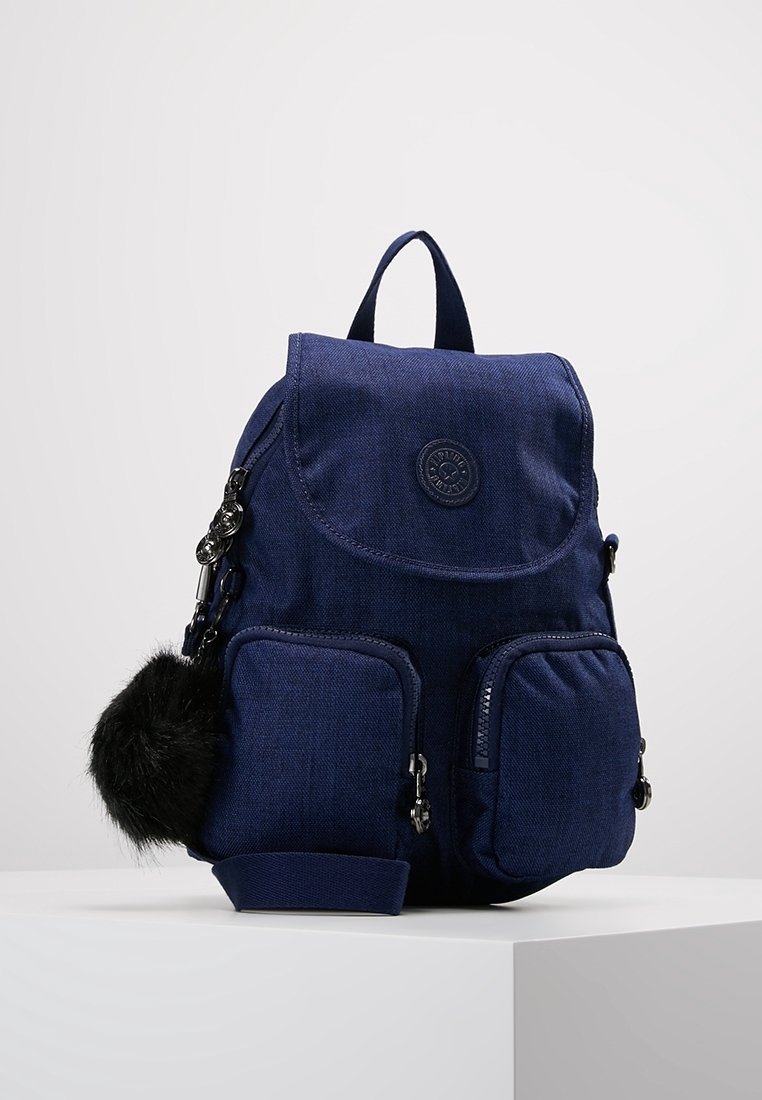 Sac à dos en tissu bleu marine avec une fermeture à rabat, deux poches zippées à l'avant et un logo rond. Il est orné d'un pompon en fourrure sur le côté.