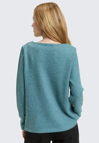 Vrouw met lang blond haar, gekleed in een teal gekleurde, gestructureerde trui met lange mouwen en zwarte broek, met haar rug naar de camera tegen een effen achtergrond.