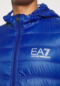 Blaues Steppjacke mit glänzender Oberfläche, Reißverschluss vorne und angenähtem Kapuzen. Mit EA7 Emporio Armani-Logo in Weiß auf der Brust.