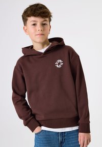 Sudadera marrón hecha de una tela suave con un bolsillo frontal tipo canguro. Presenta un logotipo circular blanco en el pecho. Se lleva sobre una camisa blanca.