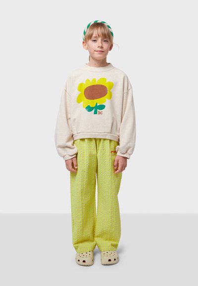 Cream sweatshirt with a large yellow flower graphic, paired with bright lime green textured pants and beige clogs. Green striped headband.