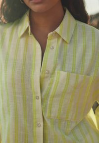 Camicia leggera con bottoni color crema a strisce verticali verdi. Caratterizzata da una tasca sul petto, colletto e texture liscia.