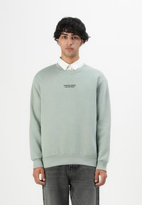 Jonge man in een lichtgroene sweatshirt met de tekst "Crafted Goods Pier One Denim," een wit overhemd eronder en vervaagde zwarte spijkerbroek.