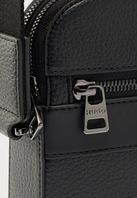 Μαύρη τσάντα από δέρμα με υφή και λείο φινίρισμα, με ασημένιο φερμουάρ με την επιγραφή "HUGO" και ανθεκτική υφασμάτινη θηλιά ιμάντα.