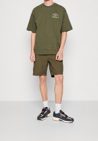 Sudadera oversize color verde oliva con logo, combinada con pantalones cortos cargo en verde a juego, y zapatillas negras/azul marino con acentos en burdeos.