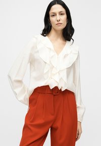 Λευκό μπλουζάκι με βολάν στο ντεκολτέ και μακριά μανίκια, συνδυασμένο με κόκκινες, πιετέ παντελόνες υψηλής μέσης. Ομαλή υφή υφάσματος.