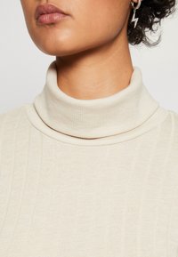 Pull à col roulé côtelé de couleur crème avec une coupe ajustée. Il présente un col haut et doux ainsi qu'une texture lisse. Accentué par une petite boucle d'oreille en forme d'éclair.