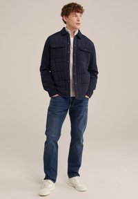 Marineblaue gesteppte Jacke mit Druckknopfverschlüssen, zwei Brusttaschen und einer lockeren Passform, kombiniert mit blauen Jeans und weißen Turnschuhen. Glatte Textur.
