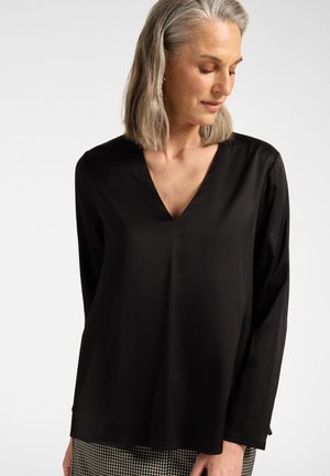 Vrouw met schouderlang grijs haar, draagt een zwarte blouse met lange mouwen en V-hals en een geruite broek, kijkt naar beneden naar de zijkant.