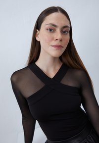 Juoda ilgomis rankovėmis palaidinė su permatomu akių viršumi, kryžminiu dizainu ir aptempta forma. Lygi tekstūra su minkštu siluetu.