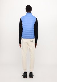 Světle modrá nadýchaná vesta s vertikálními švy, vysokým límcem, nošená přes černou košili s dlouhým rukávem. Bílé tepláky s logo detailem na zadní straně.