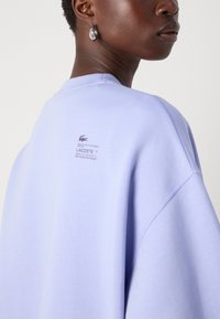 Lilla oversized genser med rund hals, med et logomerke på baksiden. Myk tekstur, avslappet passform og senket skuldersømmer.
