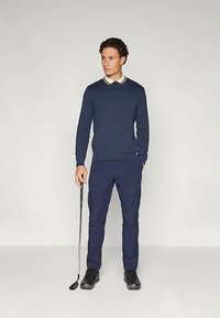 Ναυτικό πούλοβερ με σταυρωτό γιακά, συνδυασμένο με ναυτικές παντελόνες. Το παντελόνι έχει στενή εφαρμογή, και το μοντέλο κρατά έναν γκολφ κλαμπ.