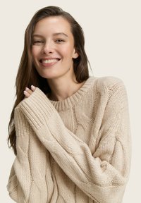 Pull en maille torsadée crème avec un col rond et des manches surdimensionnées. La texture est épaisse, reflétant un design confortable et chaud.