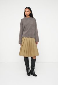 Brown knitted sweater with a round neckline, pleated beige skirt, and knee-high black leather boots, standing against a plain backdrop.