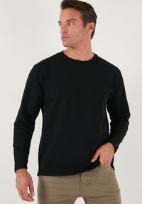 Maglietta nera a maniche lunghe con collo rotondo, realizzata in tessuto morbido, indossata sopra pantaloni chiari. Presenta un taglio dritto e nessun motivo visibile.