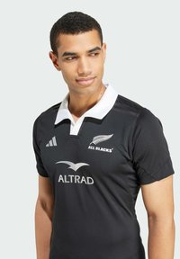 Maglia da rugby nera realizzata in tessuto leggero, con colletto bianco. Include i loghi "All Blacks" e "ALTRAD" in colori contrastanti.