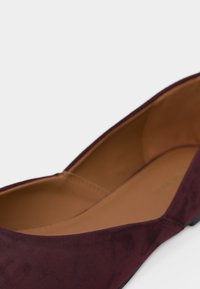 Κοντινό πλάνο ενός μοβ σκούρου σουέντ πλατ παπουτσιού με ταμπά δερμάτινη εσωτερική σόλα και χαμηλό μαύρο τακούνι σε λευκό φόντο.