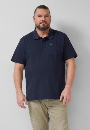 Brodaty mężczyzna stojący w ciemnoniebieskiej koszulce polo z krótkim rękawem i beżowych spodniach na jednolitym jasnoszarym tle.