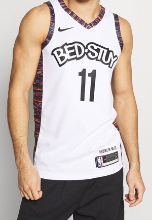 Mężczyzna noszący białą koszulkę do koszykówki Brooklyn Nets z napisem „BED-STUY” i numerem 11, z wielokolorowymi lamówkami i czarnymi szortami.