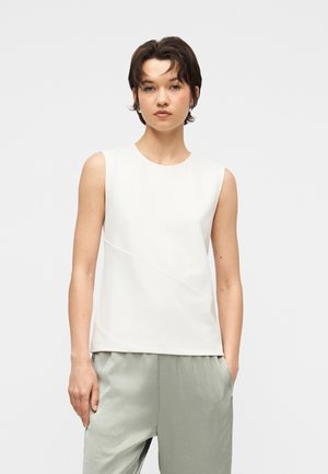 Ung person med kort mørkt hår iført en ærmeløs hvid top og lysegrå bukser, stående foran en ensfarvet baggrund.