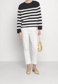 Modrý a bílý pruhovaný svetr s červeným logem, v kombinaci s bílými úzkými kalhotami a žlutými botami na bloku s podpatkem, držením malé žluté tašky.