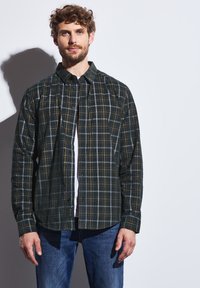 Tmavě zelená kostkovaná košile se žlutými a bílými akcenty. Má zapínání na knoflíky vpředu a dlouhé rukávy, nošená přes bílou košili. Ležérní střih.