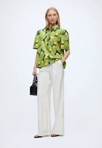 Femme portant une chemise verte à manches courtes à fleurs, pantalon blanc à jambes larges, sandales noires, tenant un petit sac à main noir, debout devant un fond blanc.