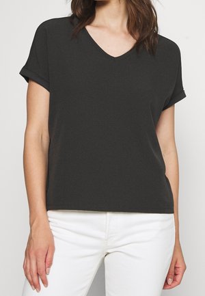 Kvinde iført en sort V-hals t-shirt med korte ærmer og hvide bukser, stående mod en ensfarvet lys baggrund.