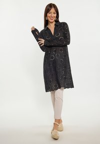 Rochie tunică neagră cu decupaje circulare, guler și tiv ondulat, asociată cu pantaloni bej deschis și sandale țesute.