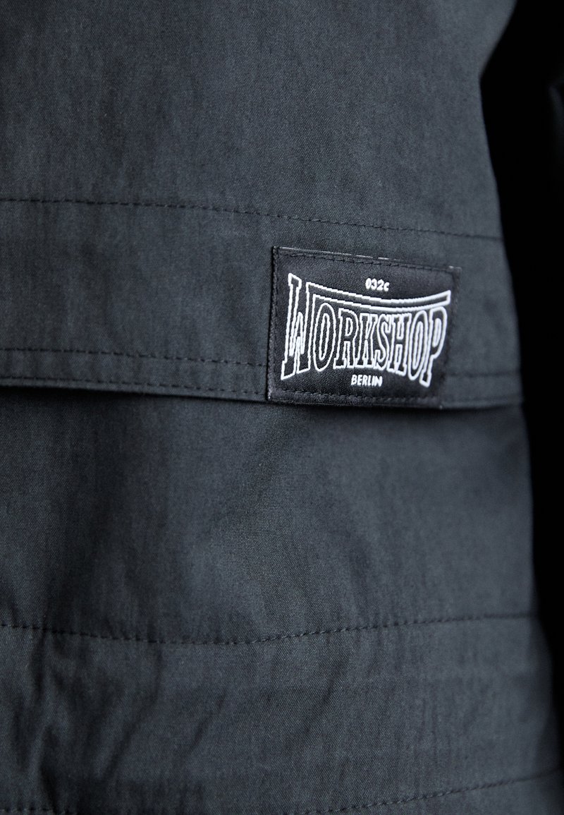 Chaqueta de tela negra con una etiqueta cosida que dice "WORKSHOP BERLIN" en blanco. El material tiene una textura suave con detalles de costura sutiles.