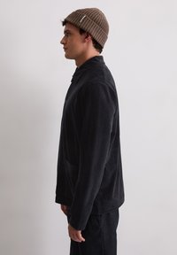 Zwarte, zachte stoffen jas met een kraag, knopen aan de voorkant en zijzak; gecombineerd met een geribbelde, bruine gebreide beanie.