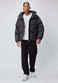 Doudoune noire avec capuche, texture matelassée, poches avant et boutons-pression, portée sur un t-shirt blanc et un pantalon noir.