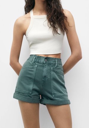 Kvinde iført hvid halter-croptop og grønne højtaljede, opsmøgede shorts med frontlommer mod en ensfarvet baggrund.