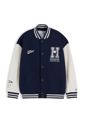 Dreng med lysbrunt hår iført navy og hvid varsity-jakke og blå jeans med trykte mønstre, står mod en ensfarvet hvid baggrund.