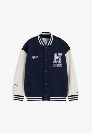 Dreng med lysbrunt hår iført navy og hvid varsity-jakke og blå jeans med trykte mønstre, står mod en ensfarvet hvid baggrund.