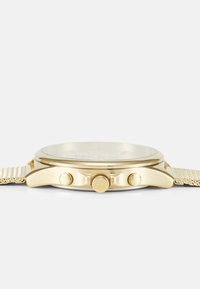 Montre en métal doré avec un boîtier rond, un bracelet en maille texturé, trois boutons-poussoirs et un verre clair mettant en valeur un cadran minimaliste.