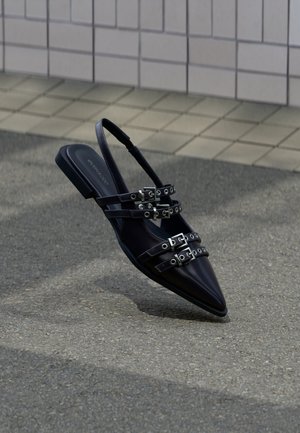 Sandalo basso nero con punta a punta e più cinturini con fibbia, con cinturino posteriore, posizionato sopra una superficie pavimentata e con sfondo di muro piastrellato.