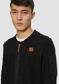 Zwarte zip-up sweater met ribbelstructuur, voorzien van een logo patch op de borst, zijzakken en een contrasterende ritslip.