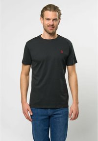 Juoda medvilninė marškinėlių su apvalia iškirpte, trumpomis rankovėmis ir maža raudona emblema kairėje krūtinės pusėje, derinama su mėlynomis džinsinėmis kelnėmis.