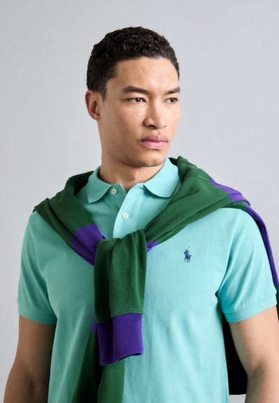 Turkos polo-shirt med korta ärmar, med en liten logotyp. En grön och lila tröja är draperad över axlarna.