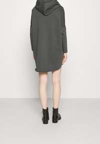 Person som bär en mörkgrå huvtröja med lång design och svarta ankelstövlar, stående barfota bakom en enkel ljus bakgrund.