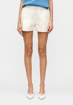 Kobieta ubrana w jasnobeżowe szorty w kwiatowy wzór z wiązaniem w pasie, niebieską bluzkę oraz białe szpilki z ozdobą z koronki na czubku.