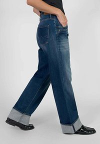 Jeans de mezclilla en azul oscuro con un ajuste holgado, puños enrollados y dos bolsillos traseros que presentan un detalle de costura de logo distintivo.