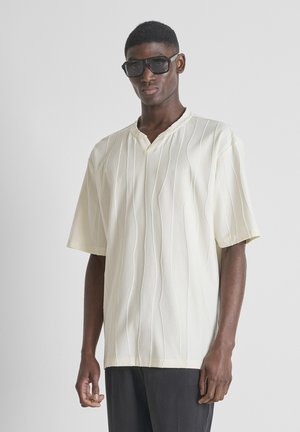 Muškarac nosi preveliku bijelu košulju kratkih rukava s vertikalnim teksturiranim prugama i velike crne sunčane naočale, stoji na jednobojnoj pozadini.