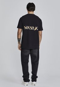 Černé bavlněné tričko s grafickým textem "SKSILK" zlatou barvou na zadní straně. Nosené s volnými tmavými džíny a bílými teniskami.