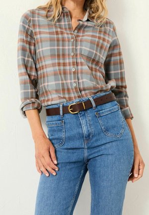 Frau trägt ein kariertes Hemd mit hochgekrempelten Ärmeln, das in eine hoch taillierte blaue Jeans gesteckt ist, dazu ein brauner Ledergürtel mit goldener Schnalle.