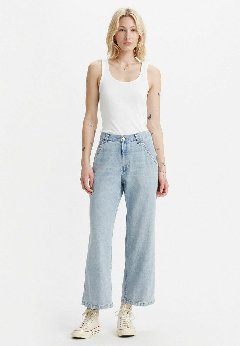 Jeans a gamba larga di un blu chiaro con vita alta, caratterizzati da una chiusura a bottone e tasche. Abbinati a una canotta bianca e sneakers beige.