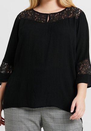 Zwarte blouse met kanten details op de halslijn en mouwen, gecombineerd met grijs-witte geruite broek, gedragen door een staand persoon met één zichtbare hand.