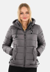 Young woman wearing a grey padded hooded jacket with zip and buttoned pockets, hands in pockets, smiling, standing against a plain background.